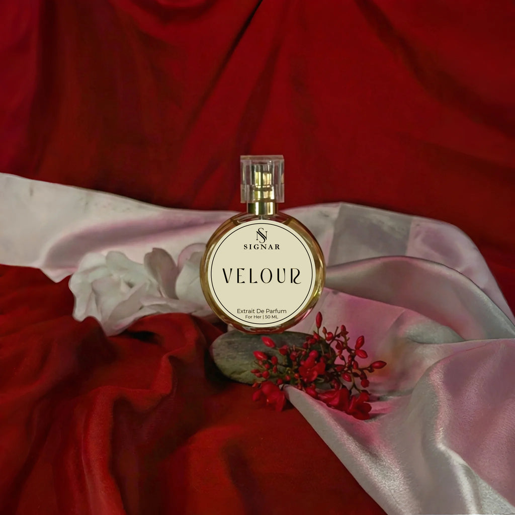 A round perfume bottle labeled 'SIGNAR VELOUR' sits on draped red and pale pink satin fabric with small red flowers around it.