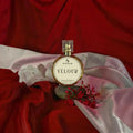 A round perfume bottle labeled 'SIGNAR VELOUR' sits on draped red and pale pink satin fabric with small red flowers around it.