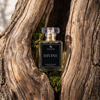 A bottle of SIGNAR Divine Extrait De Parfum placed in a hollow of weathered tree bark with small green moss visible around it. The bottle is clear glass with pale yellow liquid, a black front label with white text, and a clear cap.
