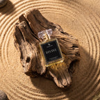 A rectangular glass perfume bottle labeled 'DIVINE' by SUGAR, placed on a piece of weathered driftwood over sand with circular ripples. The bottle contains a pale yellow liquid and has a black front label with white text.