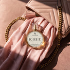 A round glass perfume bottle labeled SIGNAR ICONIC sits on draped blush pink fabric next to a matching quilted handbag with gold chain straps, in warm sunlight.