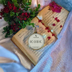 A round glass perfume bottle labeled SIGNAR ICONIC is placed on an open book with handwritten script and red petals, styled with green leaves and red flowers on draped white fabric.