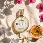 SIGNAR ICONIC Extrait De Parfum perfume bottle with a clear cap and gold spray mechanism, placed on a textured beige linen cloth with shadows, a white orchid flower, a sliced pomegranate, and pink bougainvillea flowers.