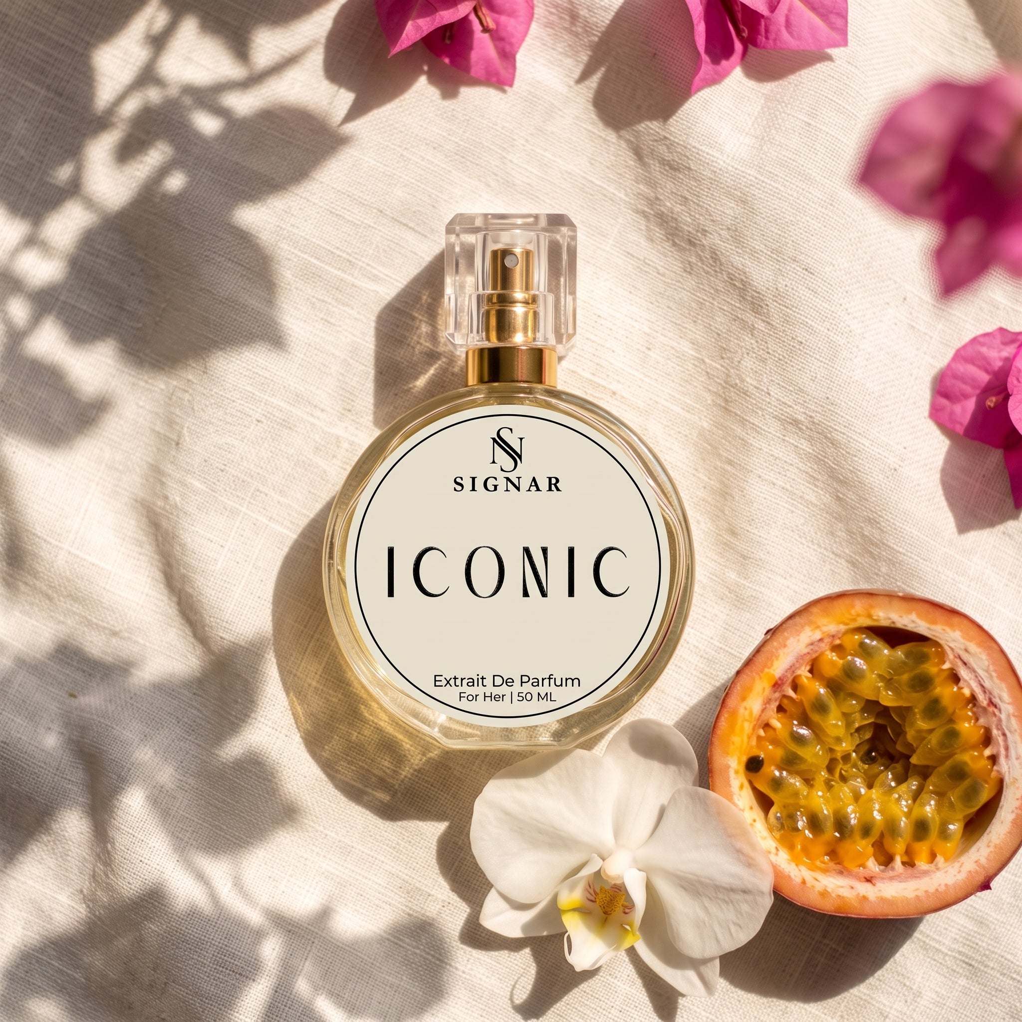 SIGNAR ICONIC Extrait De Parfum perfume bottle with a clear cap and gold spray mechanism, placed on a textured beige linen cloth with shadows, a white orchid flower, a sliced pomegranate, and pink bougainvillea flowers.