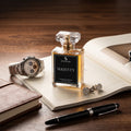 A clear glass perfume bottle labeled SIGNAR MAJESTY with amber liquid and a black label, styled on an open book with a silver watch, cufflinks, a notebook, and a black pen on a wooden table.