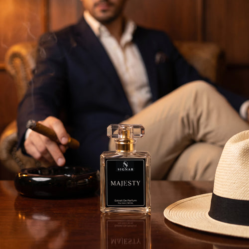 A bottle of Signar Majesty Eau De Parfum sits on a polished wooden table in a dimly lit, warm-toned lounge setting. A man in a navy blazer and beige trousers sits blurred in the background, holding a cigar next to a black ashtray.