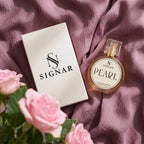 SIGNAR Pearl Extrait de Parfum perfume bottle next to its white box, styled on a draped dusty purple fabric with pink roses in the foreground.