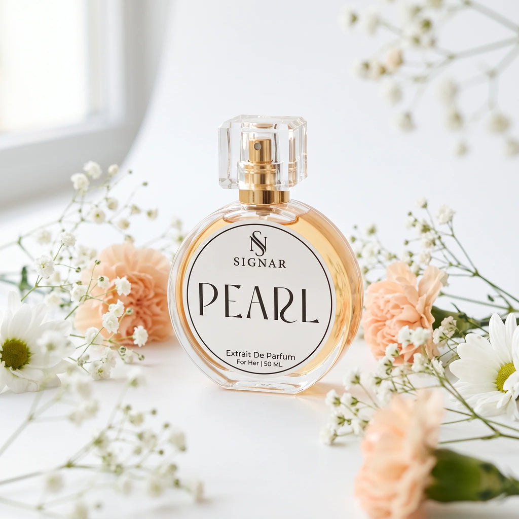 A round glass perfume bottle labeled SIGNAR PEARL with a gold spray top, styled on a white surface with soft peach flowers and white baby's breath.
