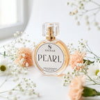 A round glass perfume bottle labeled SIGNAR PEARL with a gold spray top, styled on a white surface with soft peach flowers and white baby's breath.