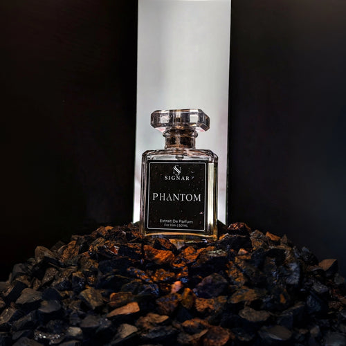A glass perfume bottle labeled 'SIGNAR PHANTOM' stands centered on a bed of dark pebbles, with a tall white illuminated backdrop behind it.