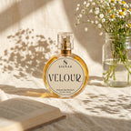 A round glass perfume bottle labeled SIGNAR VELOUR Extrait De Parfum, filled with golden liquid and topped with a clear cap, set on a sunlit cream fabric beside an open book and a glass jar of white flowers.