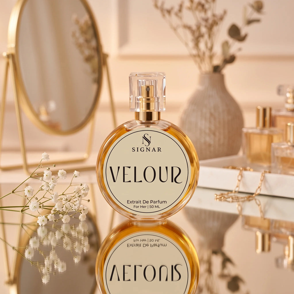 SIGNAR VELOUR Extrait De Parfum for her, 50 mL, in a round clear glass bottle with a gold spray pump and cream-colored label, displayed on a reflective surface with a gold mirror and dried flowers in the background.