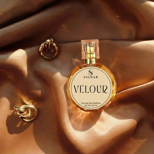 A round glass perfume bottle labeled SIGNAR VELOUR Extrait De Parfum, filled with golden liquid and topped with a clear cap, styled on draped champagne-colored satin fabric with two gold chain earrings nearby.