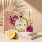 SIGNAR VELOUR Extrait De Parfum for Her, 150 mL round glass bottle with a gold cap, styled on stacked stone slabs with a lemon half, purple orchids, daisies, and a pink stone block against a warm beige background with leaf shadows.